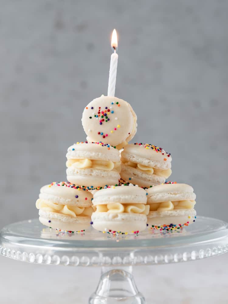 Birthday cake macarons on a platter. One has a lit birthday candle on top
