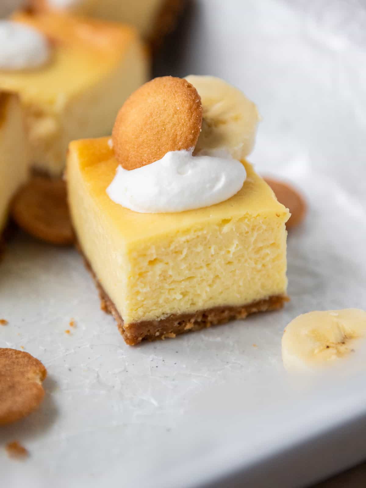 a banana pudding cheesecake bar topped with a dollop of whipped cream, a nilla wafer, and a banana slice on a white background.