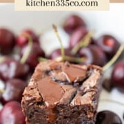 a cherry brownie sitting on top of fresh cherries and chunks of chocolate on a white background. it says cherry brownies across the top.