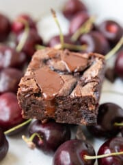 a cherry brownie sitting on top of fresh cherries and chunks of chocolate on a white background