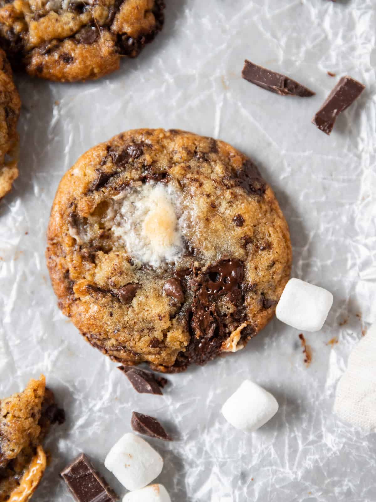 a chocolate chip marshmallow cookie surrounded by mini marshmallows and chunks of chocolate on a white background