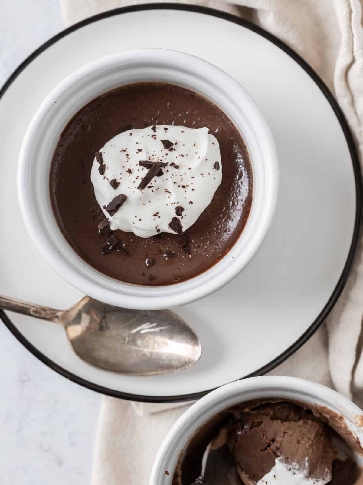a chocolate pots de crème in a ramekin with a scoop of whipped cream on top and chocolate shavings on a white plate