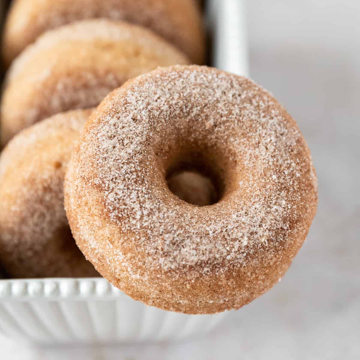 a cinnamon sugar donut balanced on the edge of a white dish with other donuts in the background