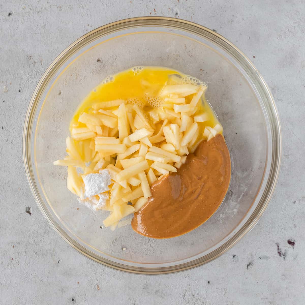 The chopped apple, peanut butter, egg, and baking powder in a glass bowl before being combined