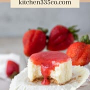 a mini strawberry cheesecake with a bite out of it surrounded by fresh strawberries on a glass dish. it says mini strawberry cheesecake across the top