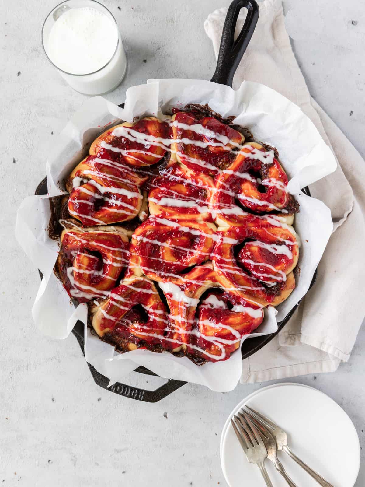 a cast iron dish filled with raspberry cinnamon rolls with a glass of milk and a dish with fork beside it
