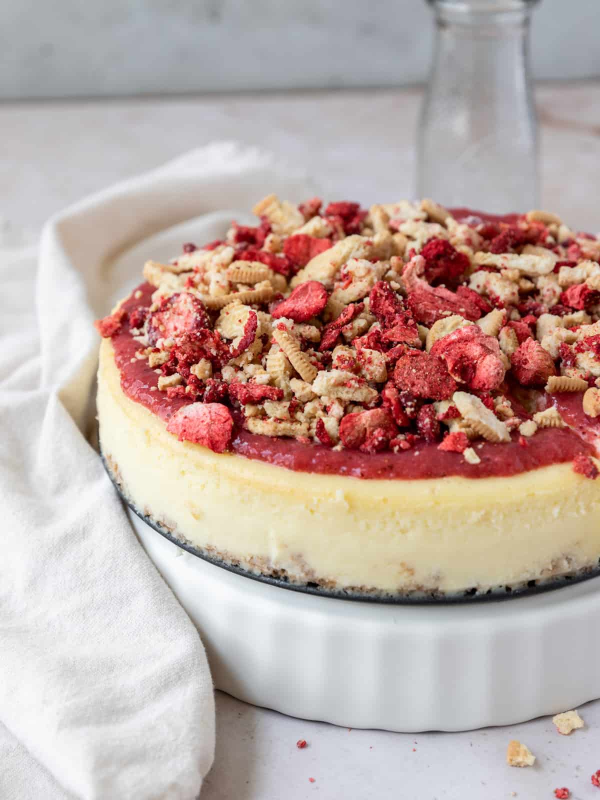a full strawberry crunch cheesecake on a white dish with a cream colored linen laying next to it