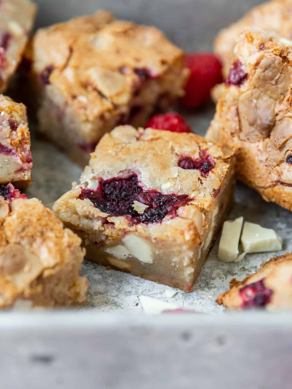 several white chocolate and raspberry blondies in a metal baking dish with chunks of white chocolate and raspberries in the bottom of the dish