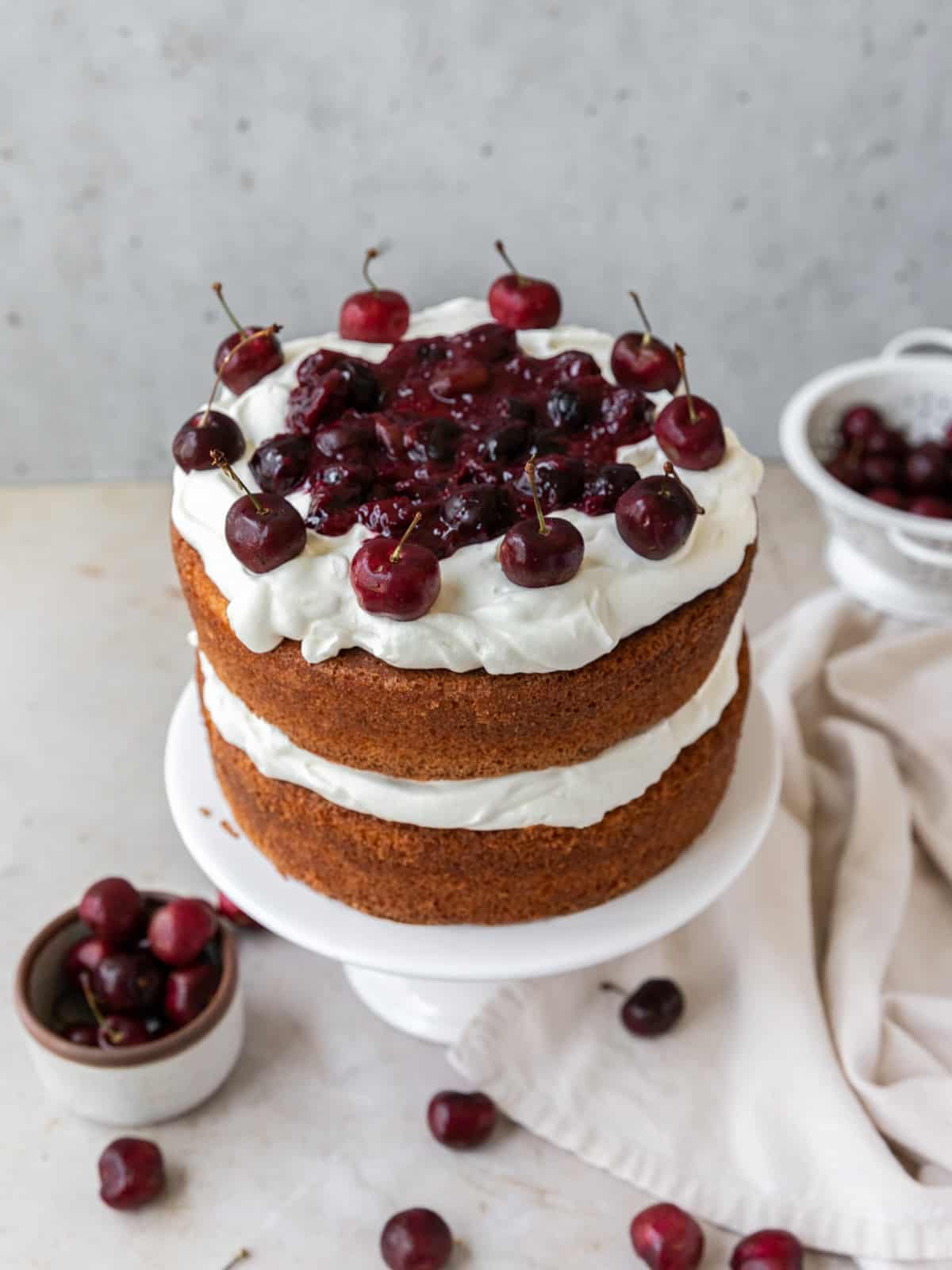 white cake layered with whipped cream and cherry compote on a white cake stand surrounded by fresh cherries