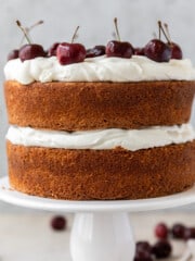 a white forest cake layered with whipped cream and cherry compote on a white cake stand surrounded by fresh cherries