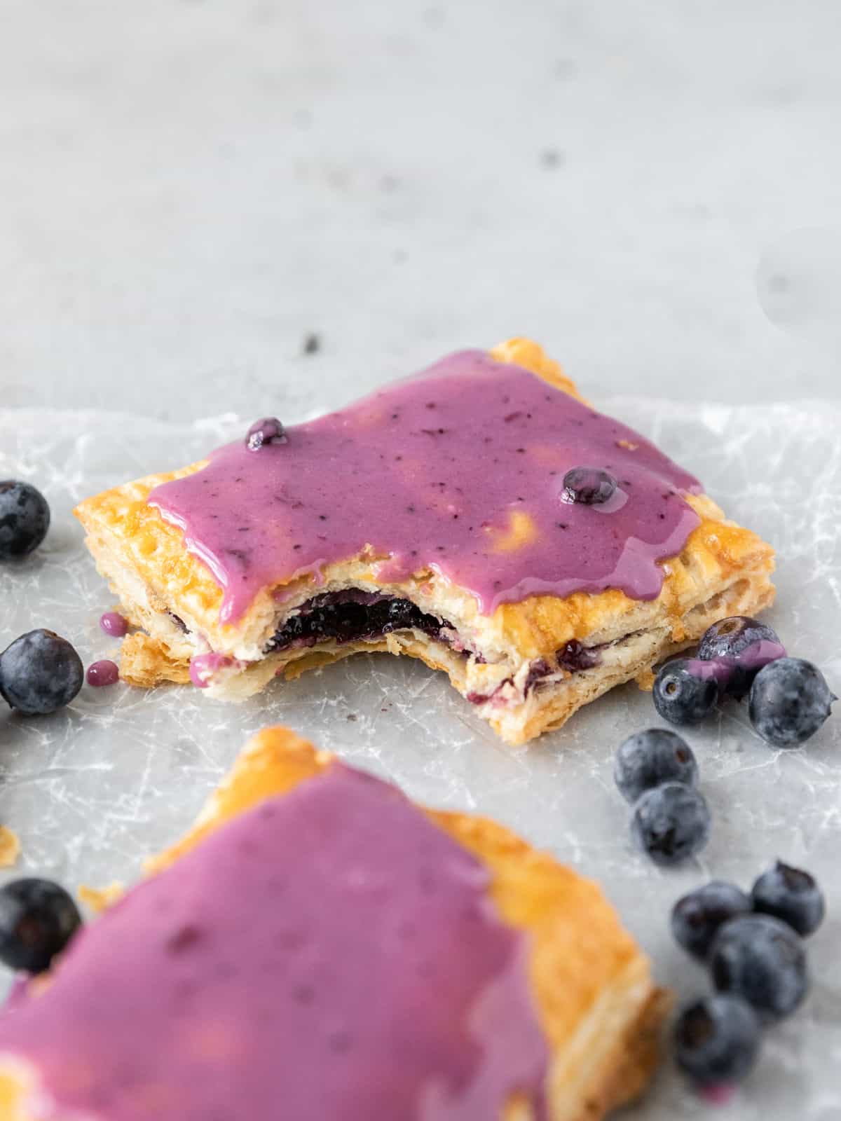 a blueberry pop tart with a bite out of it on parchment paper surrounded by fresh blueberries