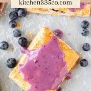 looking down on a blueberry pop tart on parchment paper surrounded by other pop tarts and fresh blueberries. It says blueberry pop tarts across the top.
