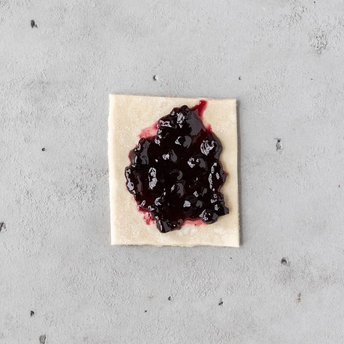 a piece of unbaked pie dough with a dollop of blueberry jam in the middle on a grey background.