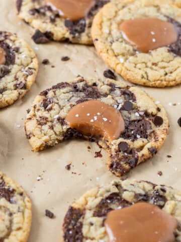 a caramel chocolate chip cookie with a bite out of it on parchment paper surrounded by other cookies.