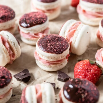 a chocolate strawberry macaron on parchment paper surrounded by other macarons, fresh strawberries, and chunks of chocolate.