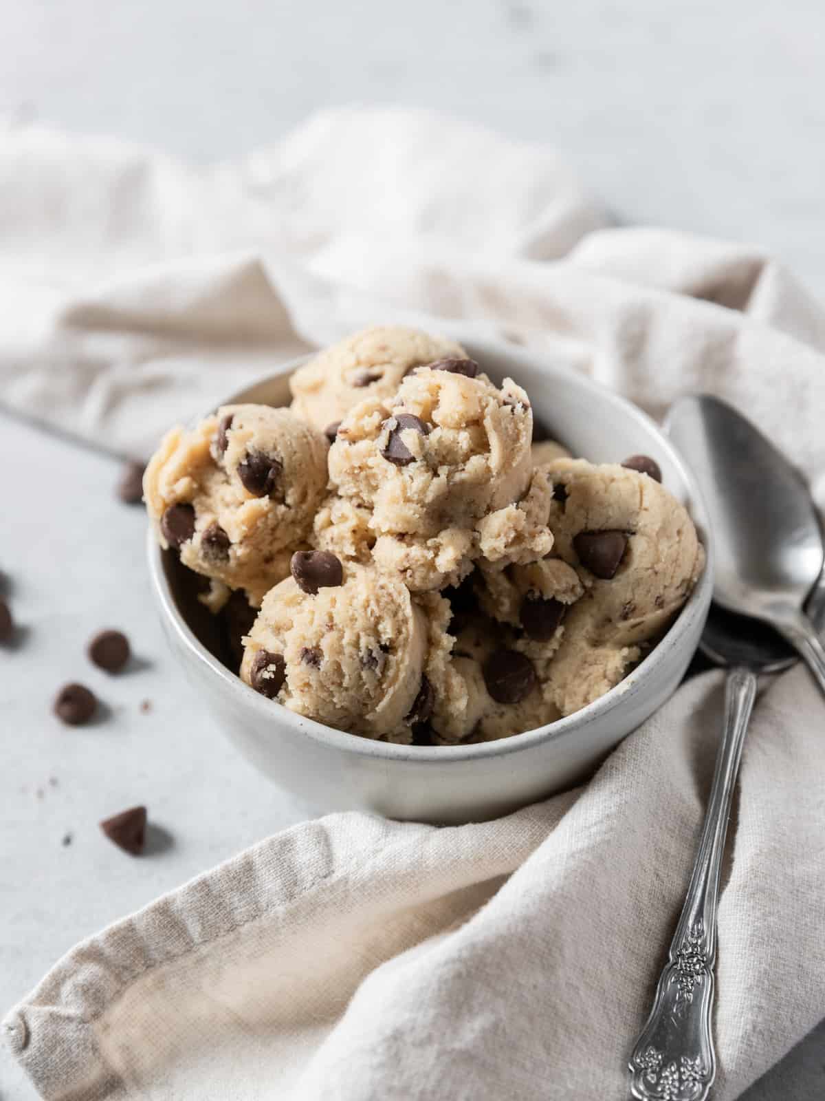 scoops of edible cookie dough in a white bowl with a linen, two spoons, and chocolate chips beside it.