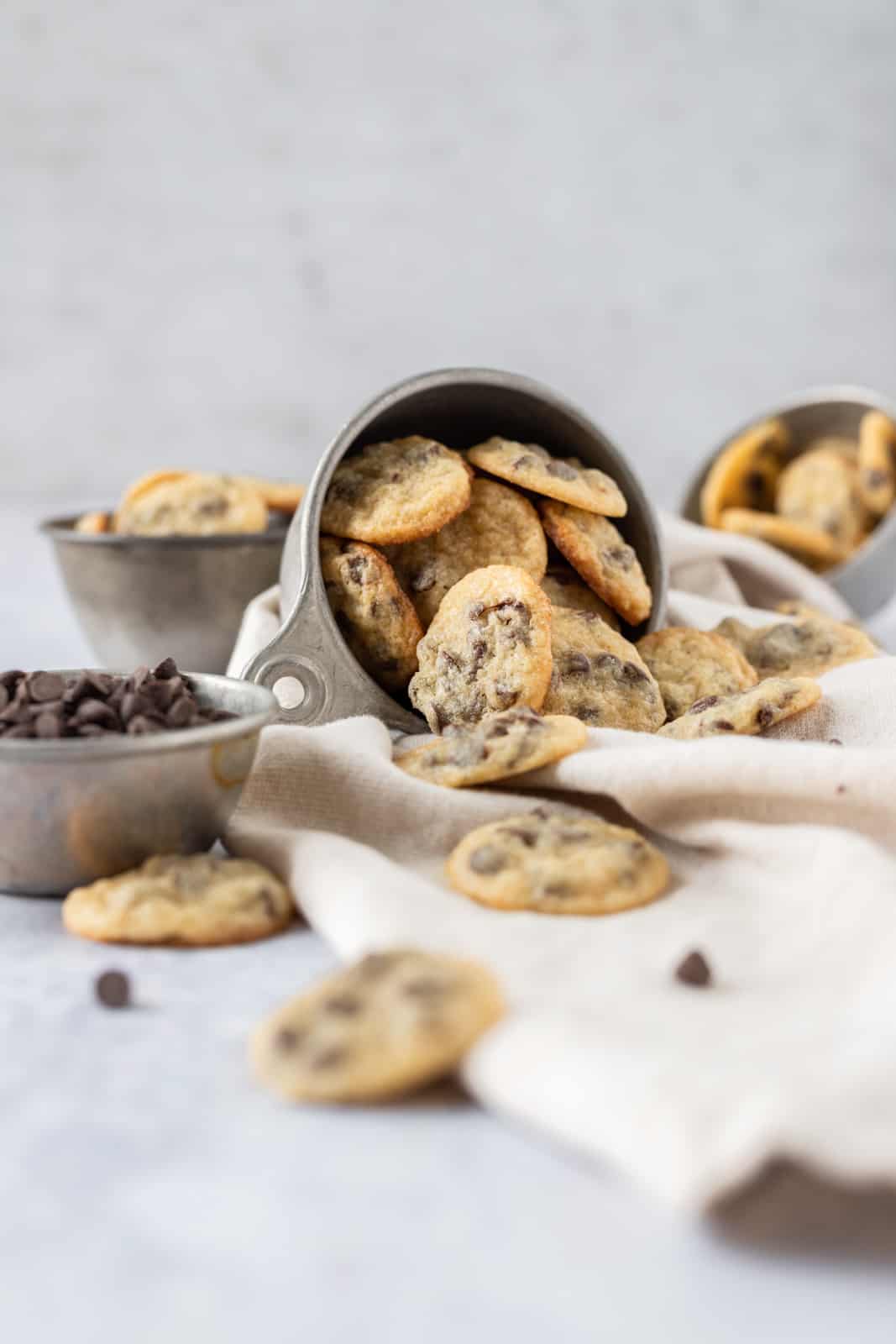 a cup full of mini chocolate chip cookies spilled over on a linen napkin.