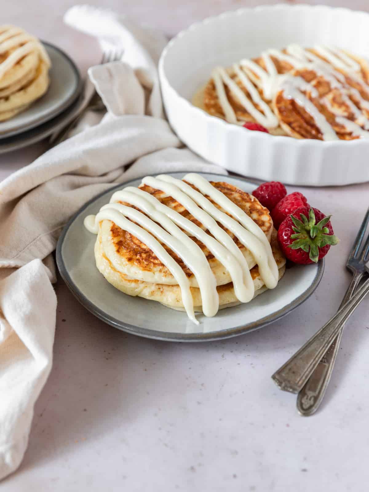 a plate of cinnamon roll pancakes with cream cheese icing there are two forks and a linen napkin next to it.