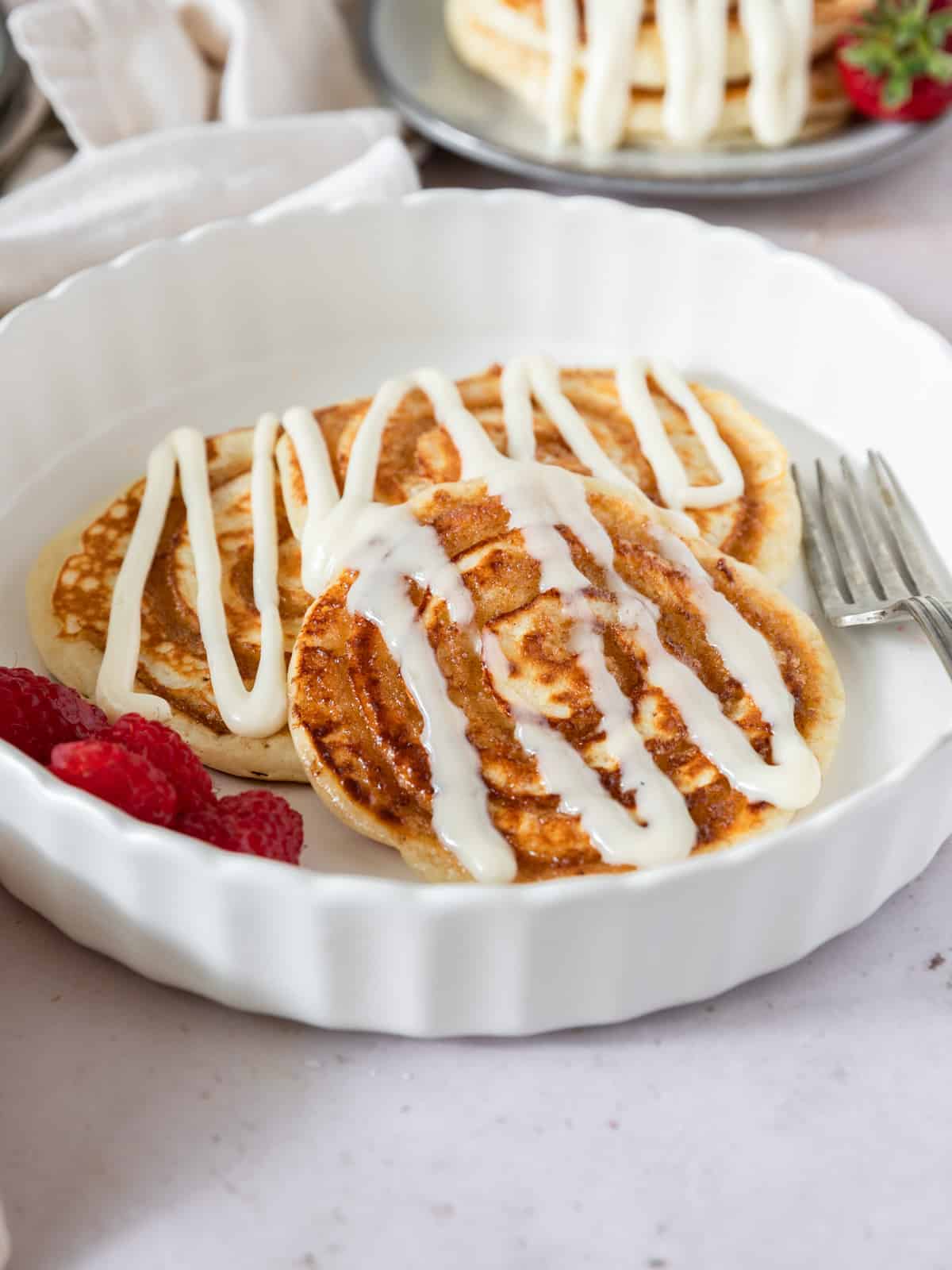 three cinnamon roll pancakes on a white plate topped with cream cheese icing.