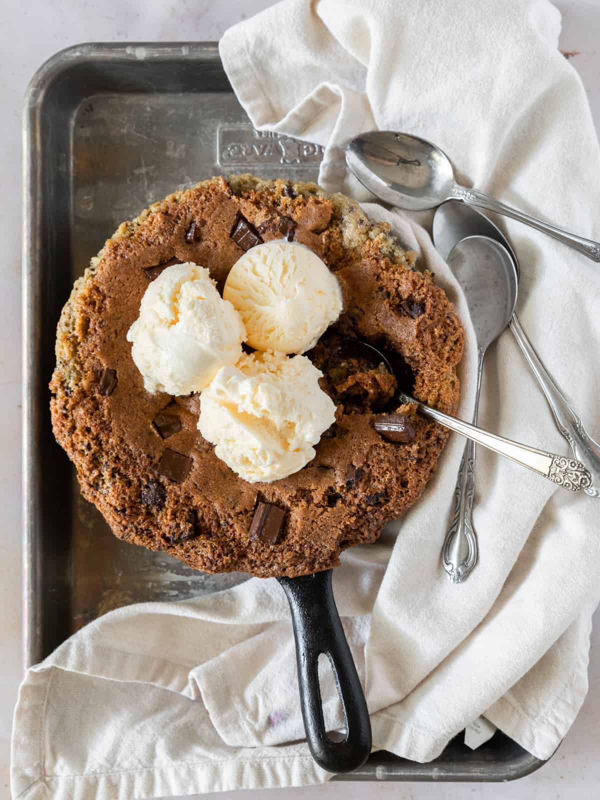 A deep dish cookie topped with three scoops of vanilla ice cream on a metal tray with a linen napkin and three spoons beside it. One spoon is taking a scoop out of it.