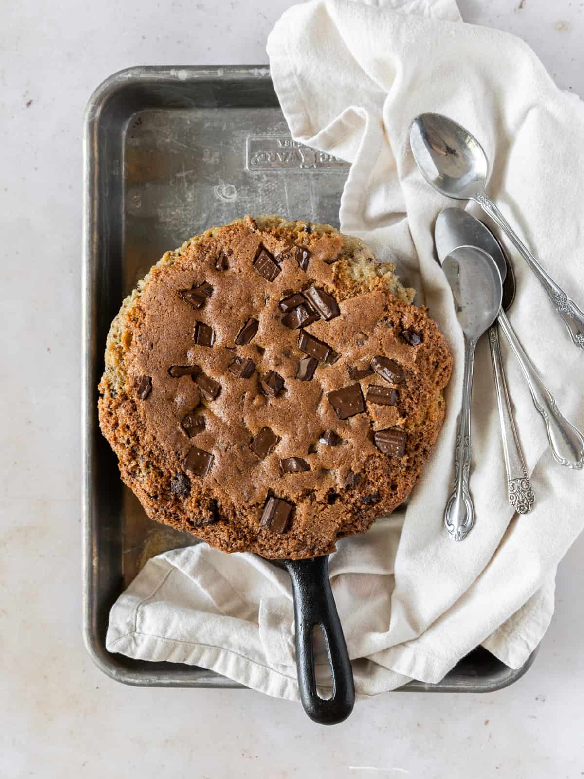 a deep dish cookie on a metal tray with a linen napkin and four spoons beside it.