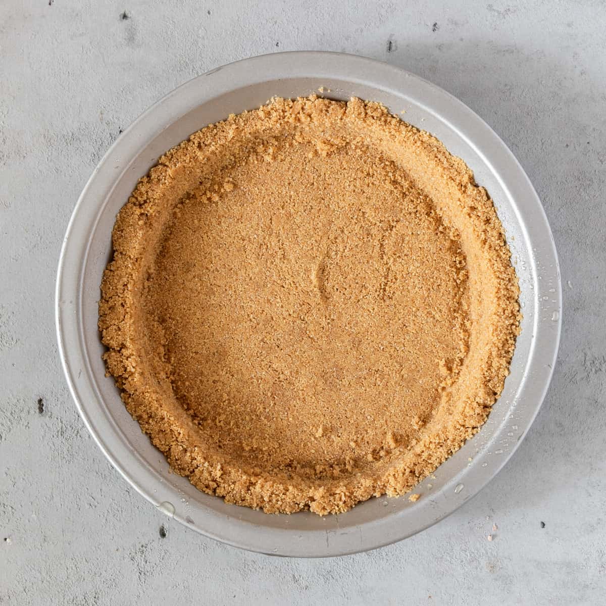 the graham cracker crust in the pie plate before being baked on a grey background.