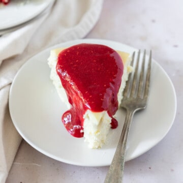 a slice of white chocolate cheesecake topped with raspberry sauce on a white plate with a fork next to it.