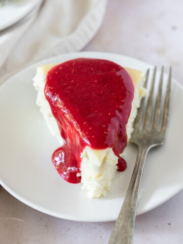 a slice of white chocolate cheesecake topped with raspberry sauce on a white plate with a fork next to it.