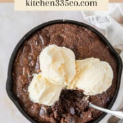 Looking down on a skillet brownie in a cast iron skillet topped with ice cream with a spoon stuck in it. It says skillet brownie across the top.