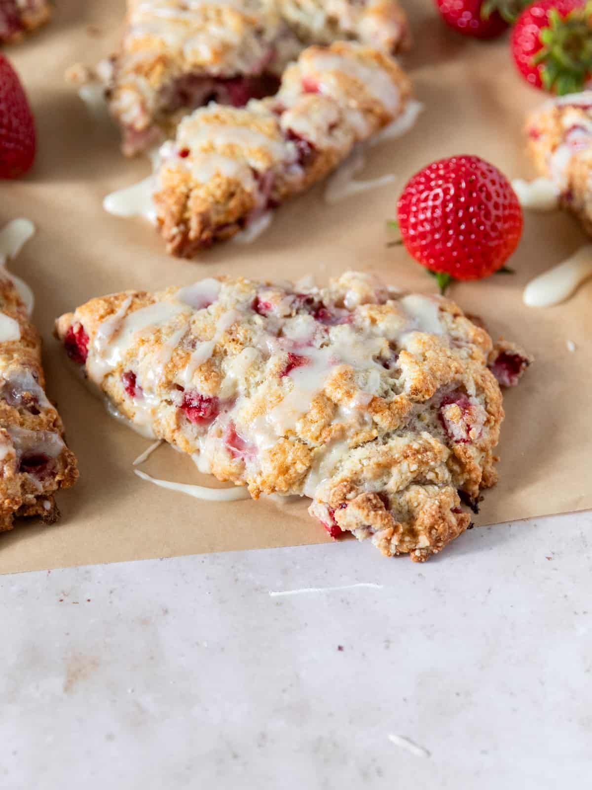 Strawberry scones on a piece of parchment paper drizzled with vanilla glaze and surrounded by fresh strawberries.