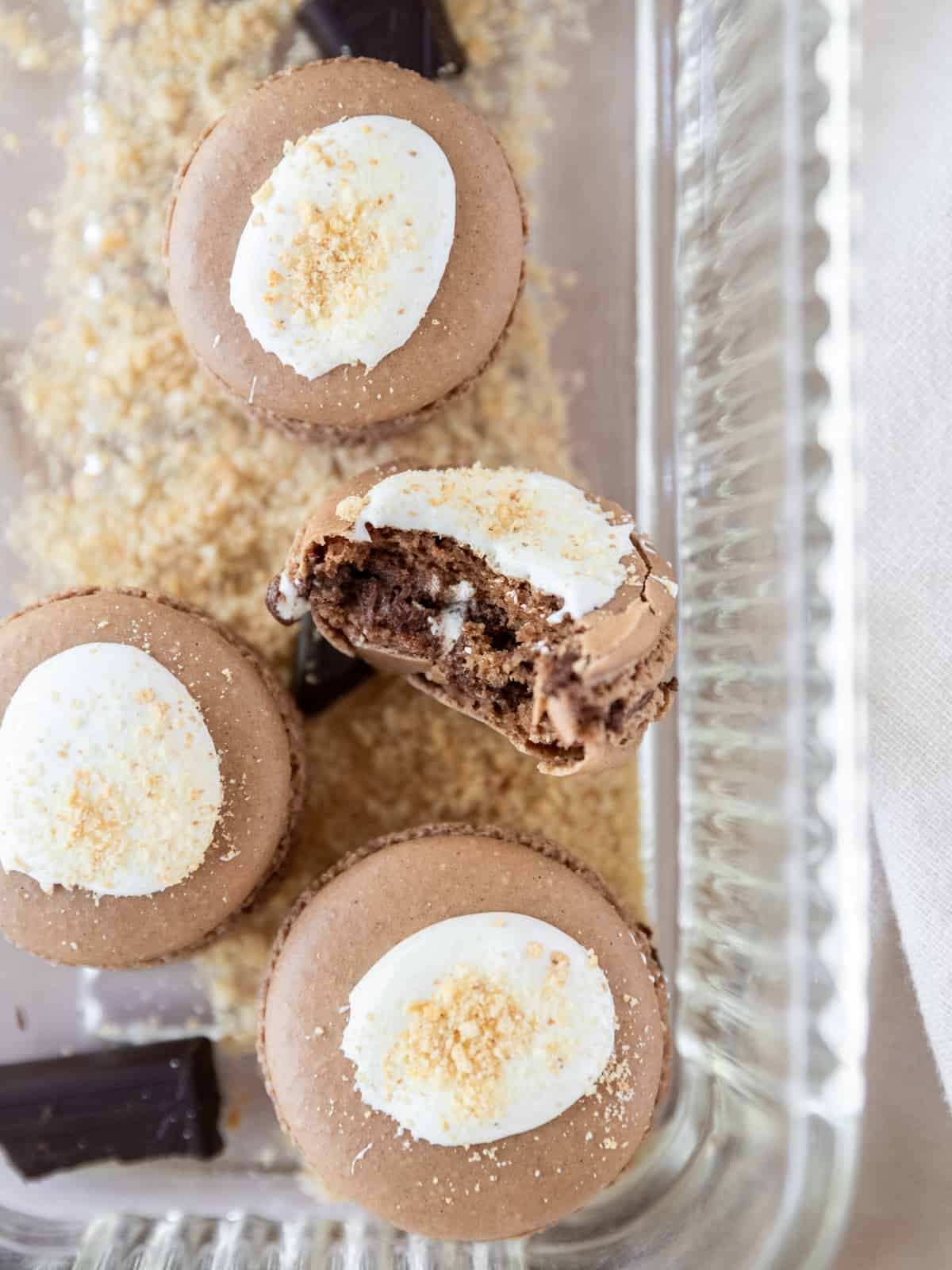 a smore macaron with a bite out of it surrounded by other macarons, chocolate, and graham cracker crumbs.