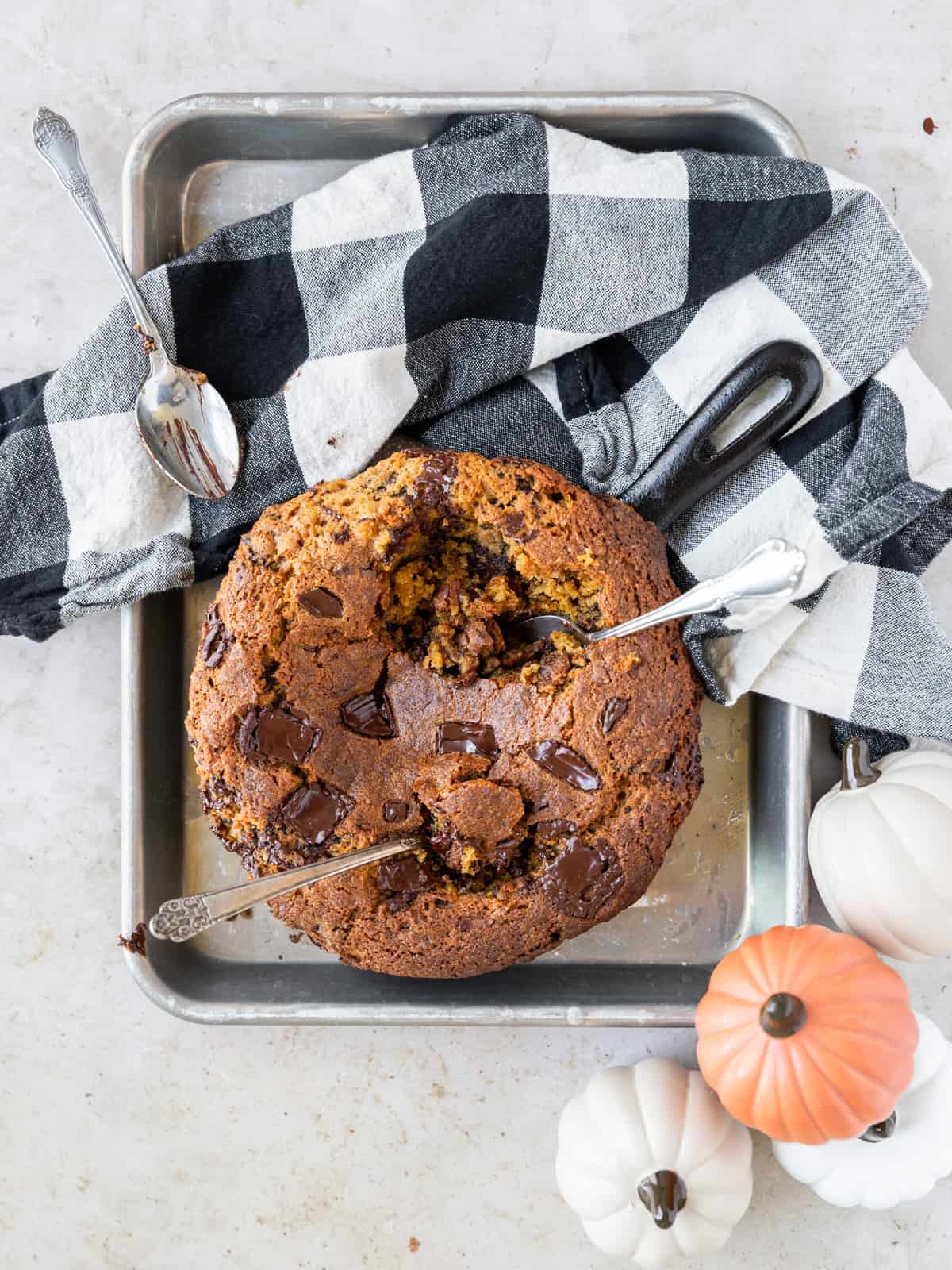 a pumpkin skillet cookie on a small metal baking pan with a black and white checked linen beside it and two spoons digging into it.