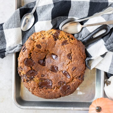 a pumpkin skillet cookie on a small baking sheet with a black and white checked linen and three spoons next to it.