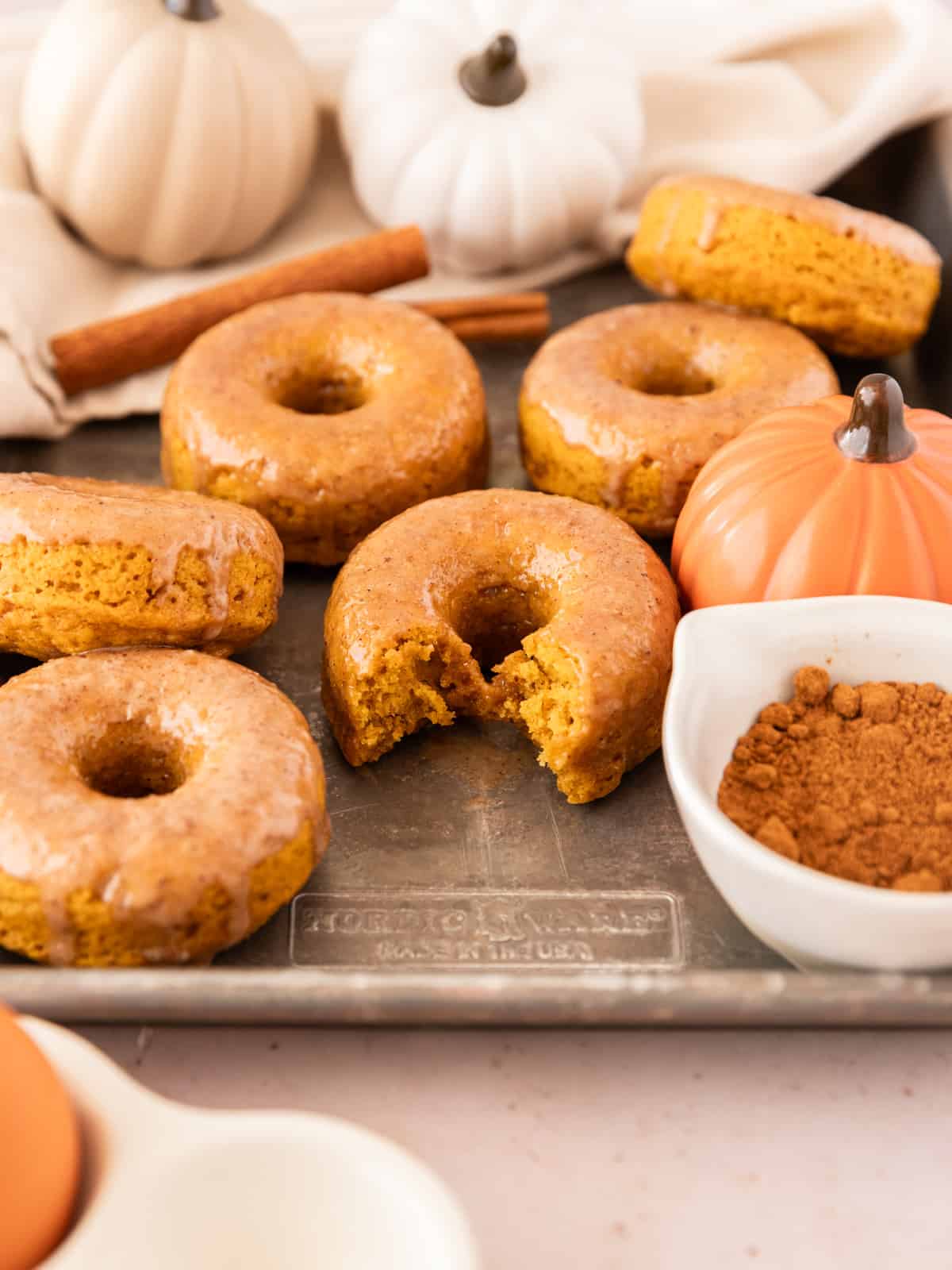 Pumpkin spice donuts on a metal sheet pan with small pumpkin and cinnamon sticks around them.
