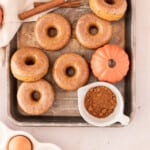 pumpkin spice donuts on a metal sheet pan surrounded by small pumpkins, cinnamon sticks, and a ramekin of pumpkin spice.