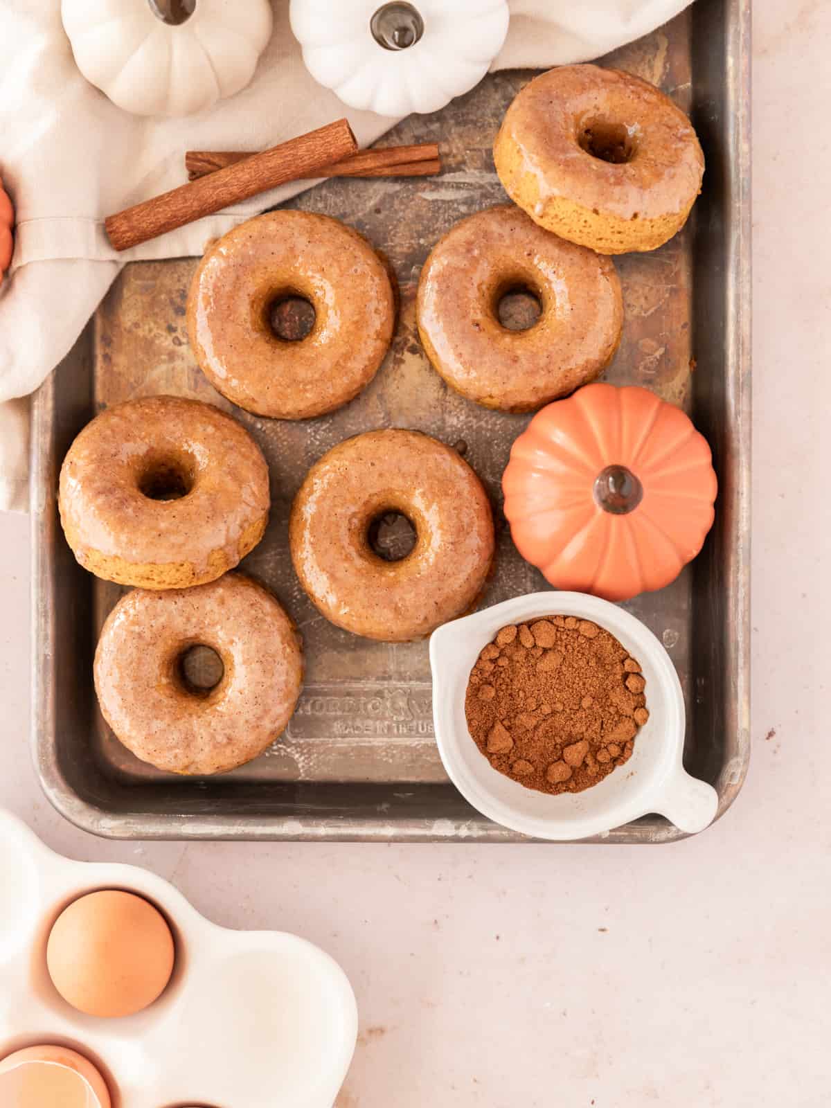 pumpkin spice donuts on a metal sheet pan surrounded by small pumpkins, cinnamon sticks, and a ramekin of pumpkin spice.