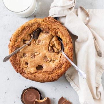 a reese's skillet cookie with two spoons in it surrounded by peanut butter cups and a glass of milk.