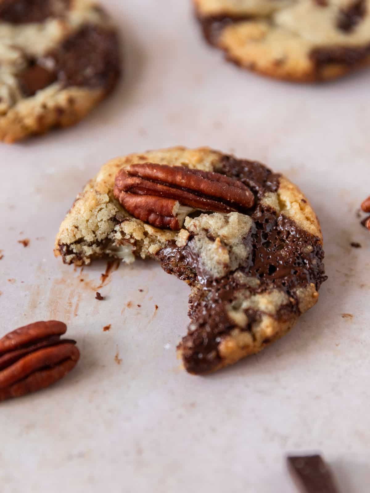 a pecan chocolate chip cookie with a bite out of it surrounded by other cookies, pecans, and chocolate.