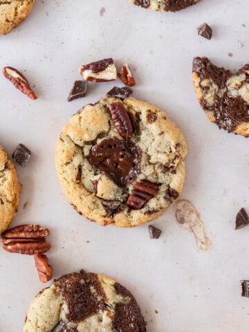 several pecan chocolate chip cookies surrounded by pecans and chunks of chocolate.