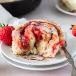 a strawberry cinnamon roll that has been cut into on a white plate with a fresh strawberry beside it.