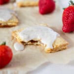 A strawberry pop-tart with a bite out of it on a piece of parchment paper surrounded by fresh strawberries.
