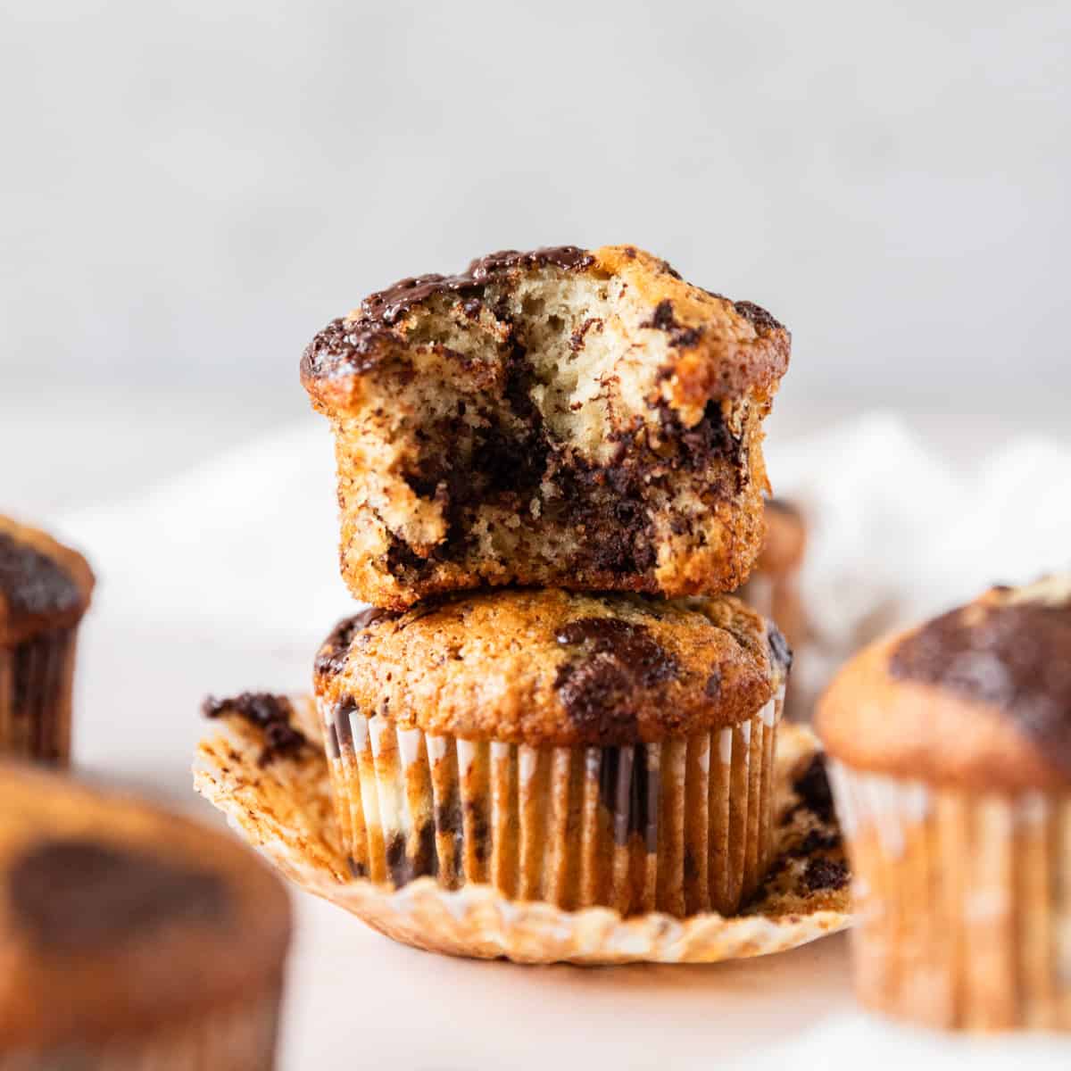 two banana chocolate chunk muffins stacked on top of each other. One has a bite out of it and there are other muffins around them.