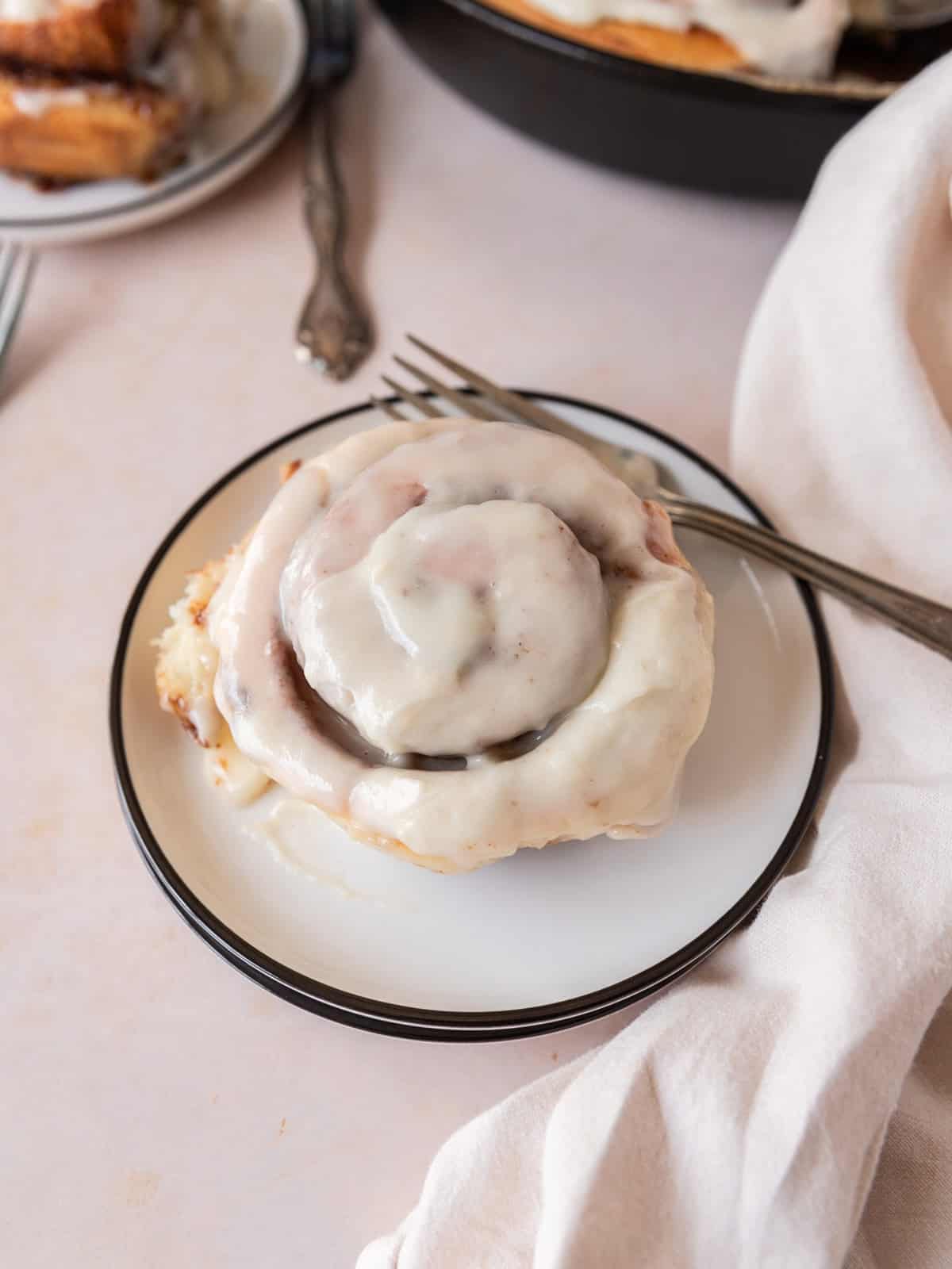 a cinnamon roll topped with icing on white plate with a metal fork next to it.