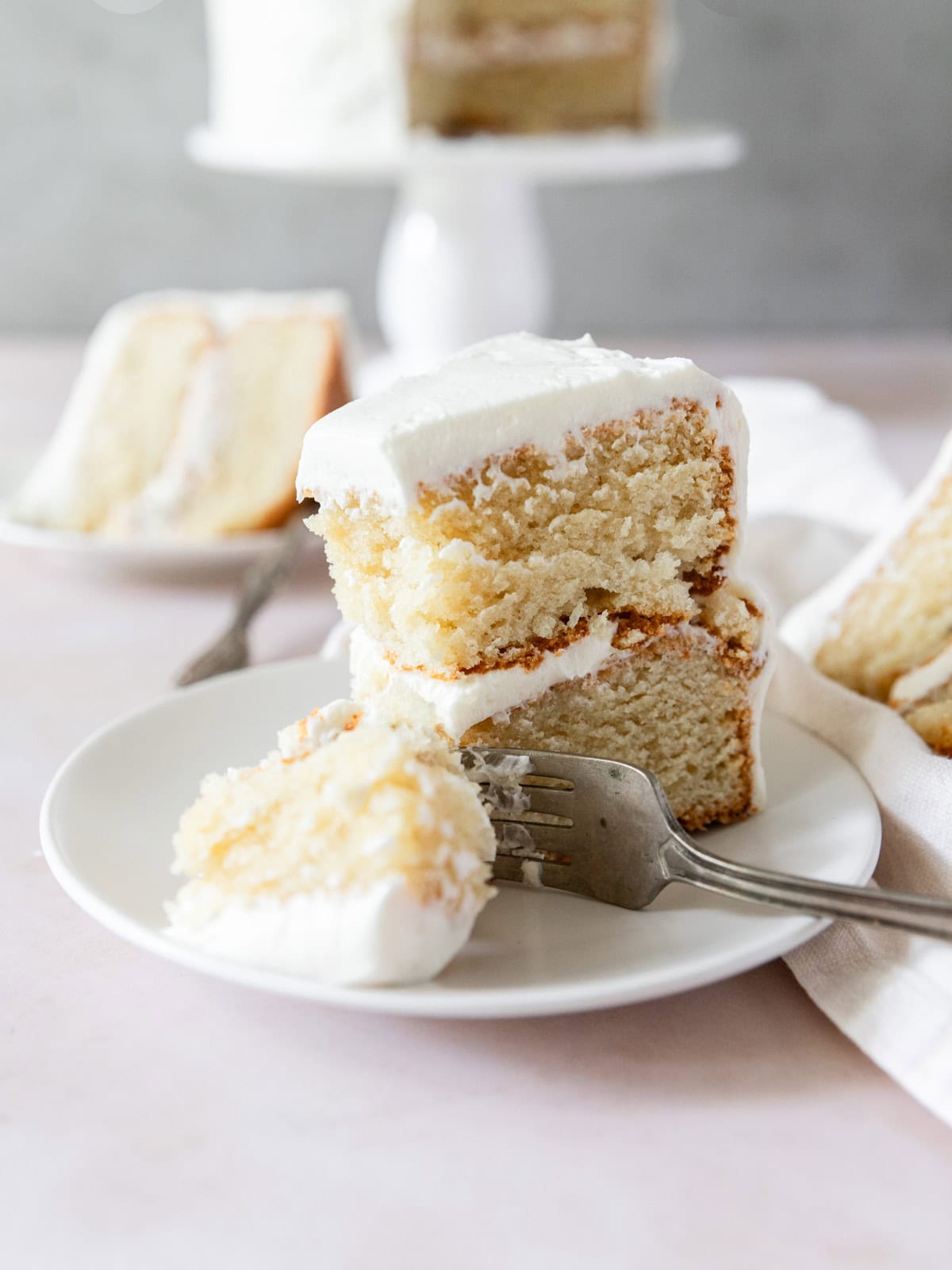 a slice of moist vanilla cake with vanilla buttercream frosting on a white plate with a bite taken out of it.