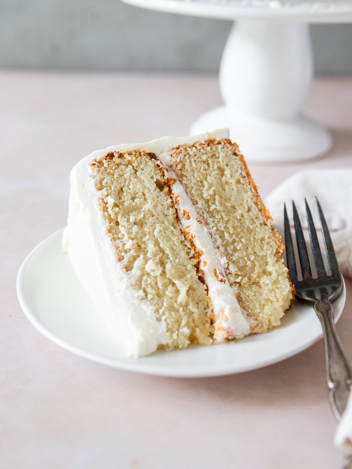 a slice of vanilla cake with buttercream frosting on its side on a white plate with a metal fork beside it.