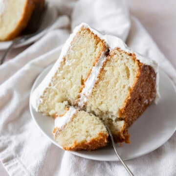 a slice of moist vanilla cake on a white plate with a bite being taken out of it.