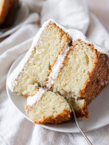 a slice of moist vanilla cake on a white plate with a bite being taken out of it.
