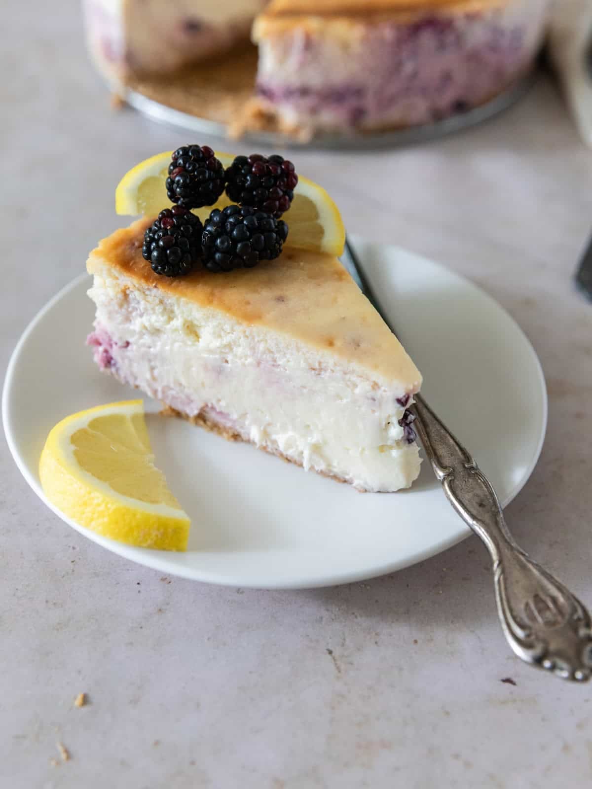 a slice of lemon blackberry cheesecake on a white plate with a metal fork. The cheesecake is topped with fresh blackberries and slices of lemon.