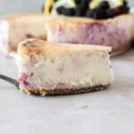 a slice of lemon blackberry cheesecake on a cake knife in front of a whole cheesecake.