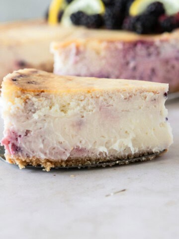a slice of lemon blackberry cheesecake on a cake knife in front of a whole cheesecake.
